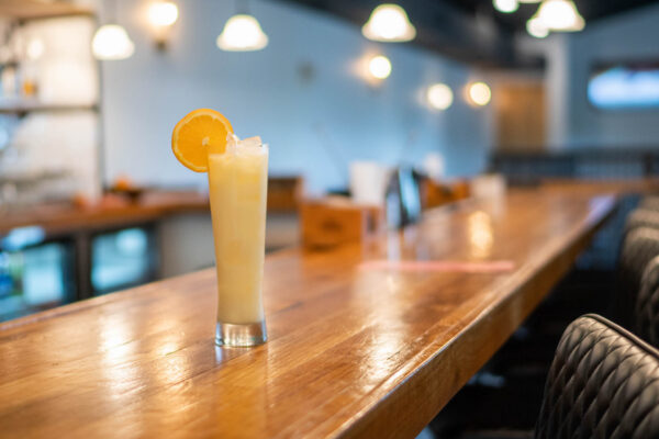 A tall glass of a creamy, iced drink garnished with an orange slice sits on a polished wooden bar counter in a softly lit, modern bar or café.