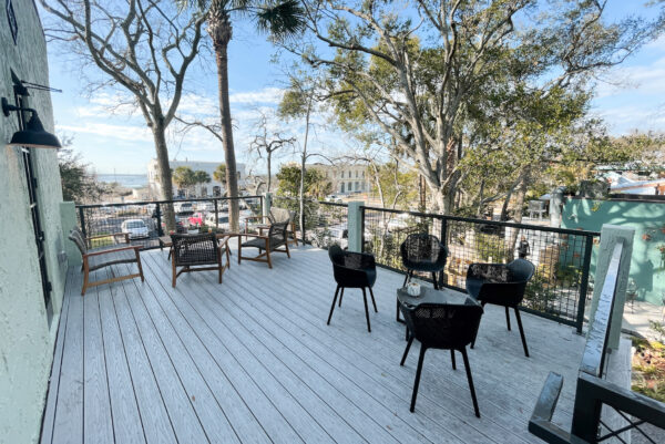 Outdoor patio with black and wooden chairs around small tables on a white wooden deck, surrounded by trees and a metal railing, overlooking a street with parked cars and distant water views.