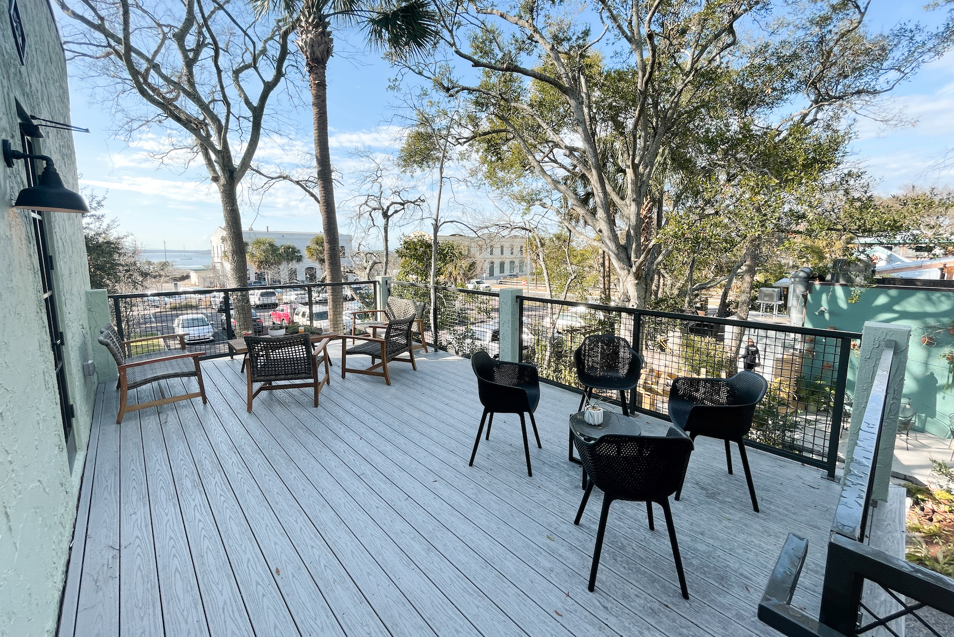 Outdoor patio with black and wooden chairs around small tables on a white wooden deck, surrounded by trees and a metal railing, overlooking a street with parked cars and distant water views.