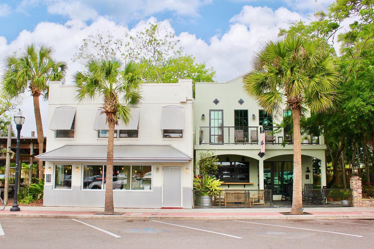 Two adjacent buildings with palm trees in front: one is white with large windows and awnings, and the other is light green with an upper balcony, both under a partly cloudy sky in a quiet, empty parking area.