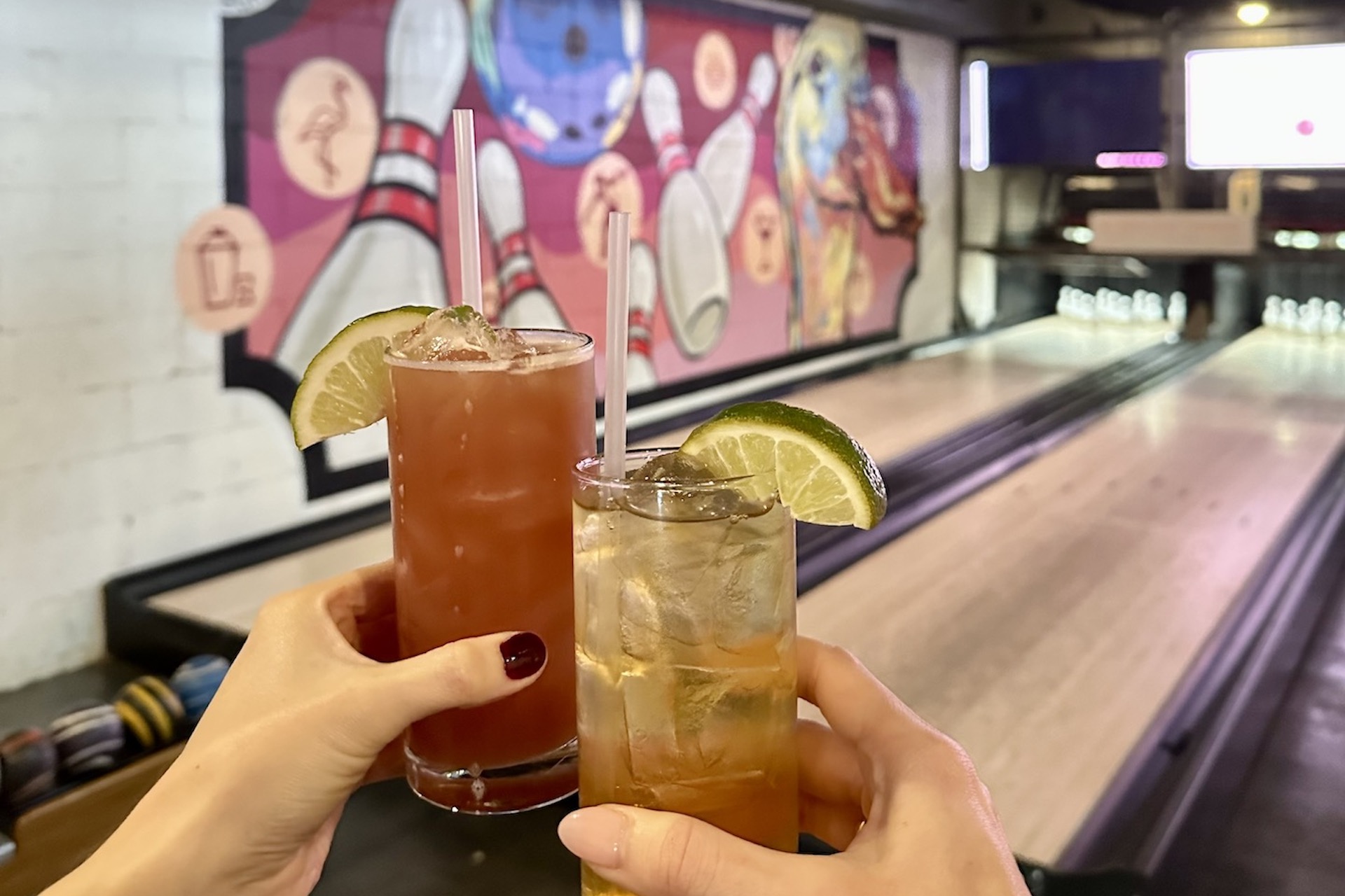 Two people holding cocktails with lime wedges, toasting at a bowling alley with colorful pins and bowling-themed mural in the background. Bowling lanes and bowling balls are visible.