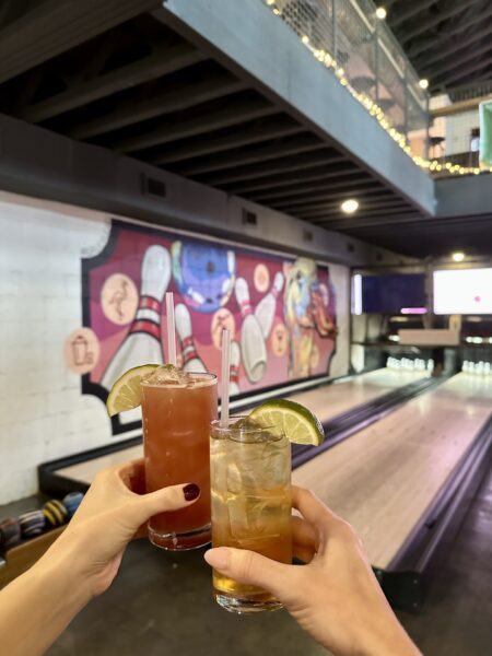 Two hands holding cocktails with lime wedges, toasting in front of a bowling lane. The background features a colorful mural of bowling pins and balls and string lights along the balcony above.