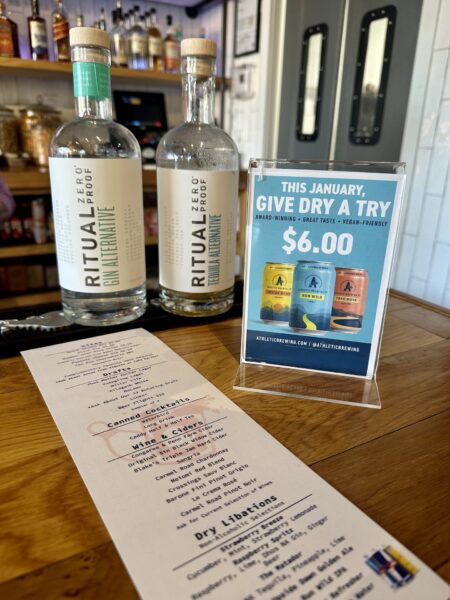 Two bottles of Ritual Zero Proof non-alcoholic spirits, a promotional sign for a $6 Dry January offer, and a cocktail menu are displayed on a wooden bar counter with shelves of bottles in the background.