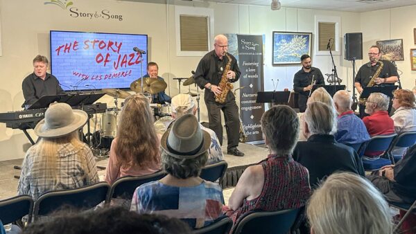 A jazz band performs on stage for an audience. A screen beside them reads The Story of Jazz with Les DeMerle. People sit attentively, some wearing hats. Musical instruments and art decorate the well-lit venue.