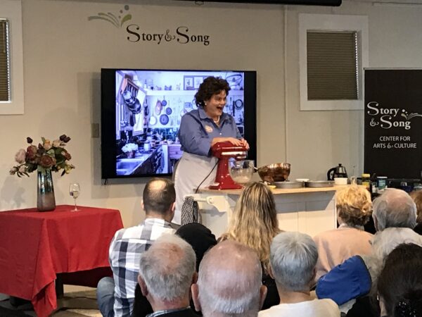 A woman in costume demonstrates cooking with a mixer on stage at the Story & Song Center for Arts & Culture, addressing an audience. A photo of a kitchen is displayed on a screen behind her.