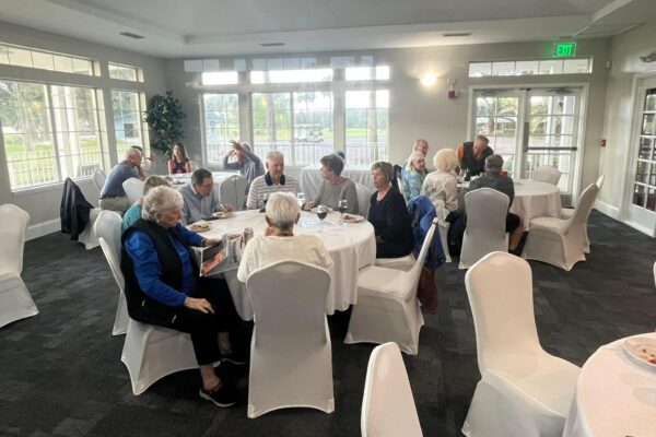 A group of older adults sit at round tables covered with white tablecloths, talking and eating in a bright room with large windows and white chairs. Some people are engaged in conversation, and natural light fills the space.
