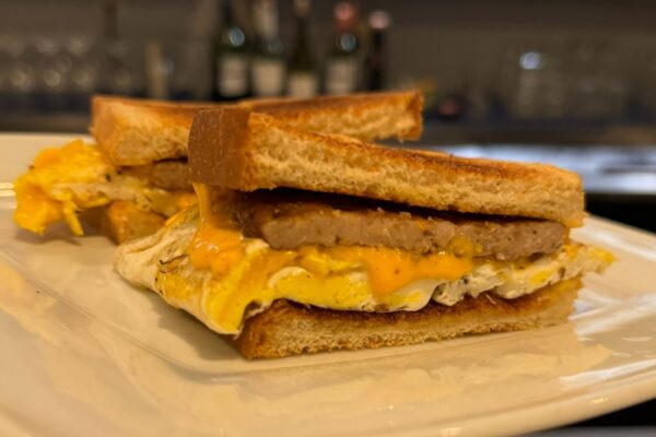 Two breakfast sandwiches made with toasted bread, eggs, melted cheddar cheese, and sausage patties are stacked on a white plate, with a blurred background of bottles and glasses.