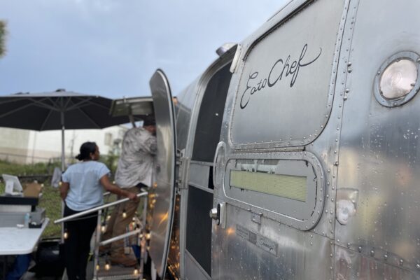 A shiny silver food truck labeled EuroChef with people standing near its open door under a large black umbrella on a cloudy day.