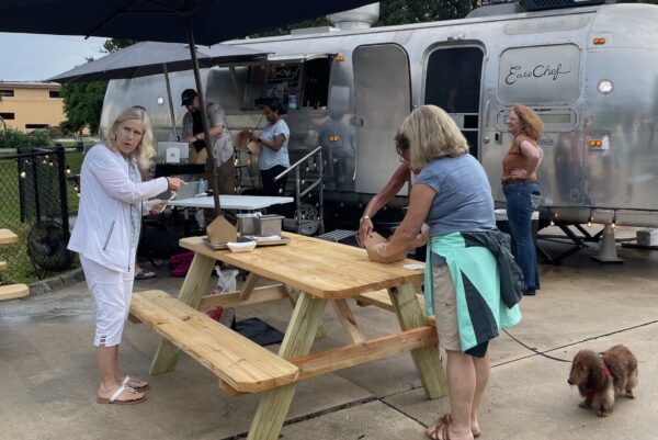 Several women stand around a wooden picnic table near a shiny food truck. One woman cleans her hands, while another holds a leash attached to a small brown dog. People are ordering food at the truck in the background.