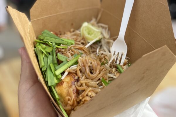 A hand holds a takeout box filled with pad thai noodles, green onions, bean sprouts, tofu, and a wedge of lime, with a white plastic fork inside.