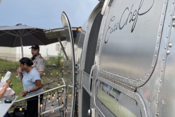 A close-up of a silver food truck with Eat Chef written on the side, parked outdoors. In the background, people are ordering and waiting near the truck under an umbrella.