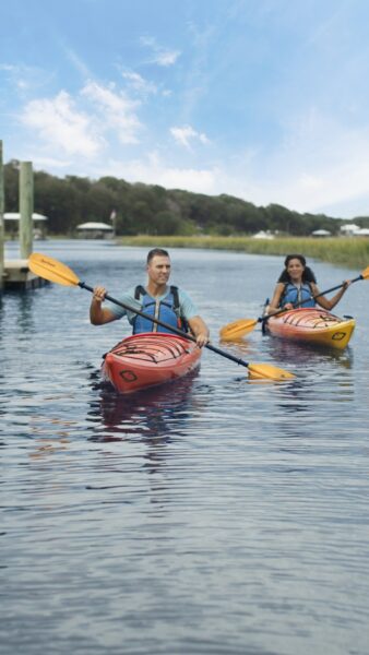 Two people paddle red kayaks on calm water near a dock, surrounded by greenery and under a bright blue sky with scattered clouds.