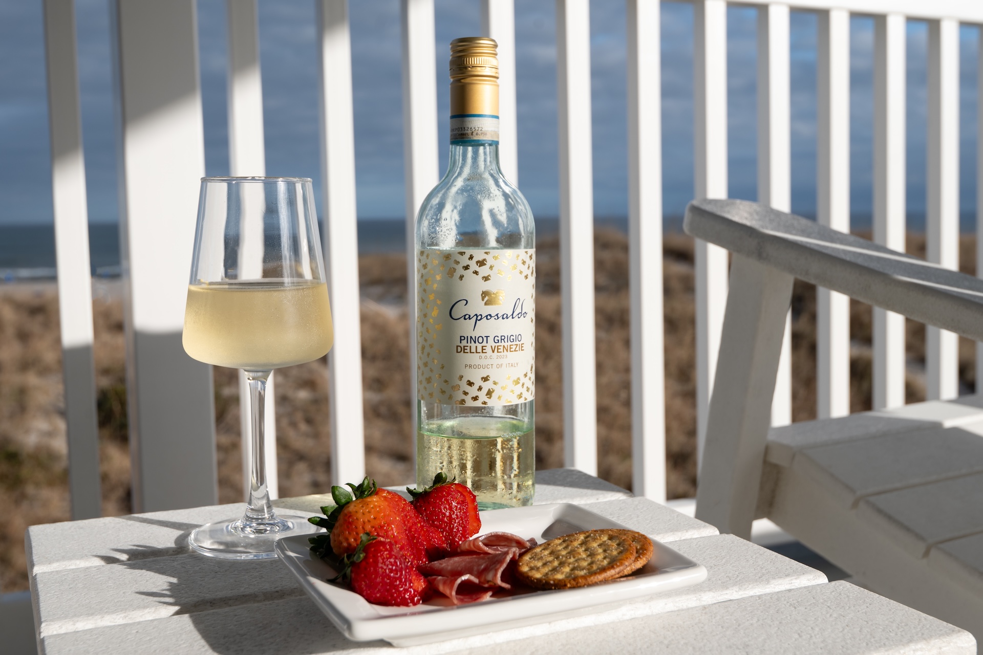 A bottle of Caposaldo Pinot Grigio, a filled wine glass, and a plate with strawberries, prosciutto, and crackers sit on a white table on a balcony overlooking sand dunes and the ocean.
