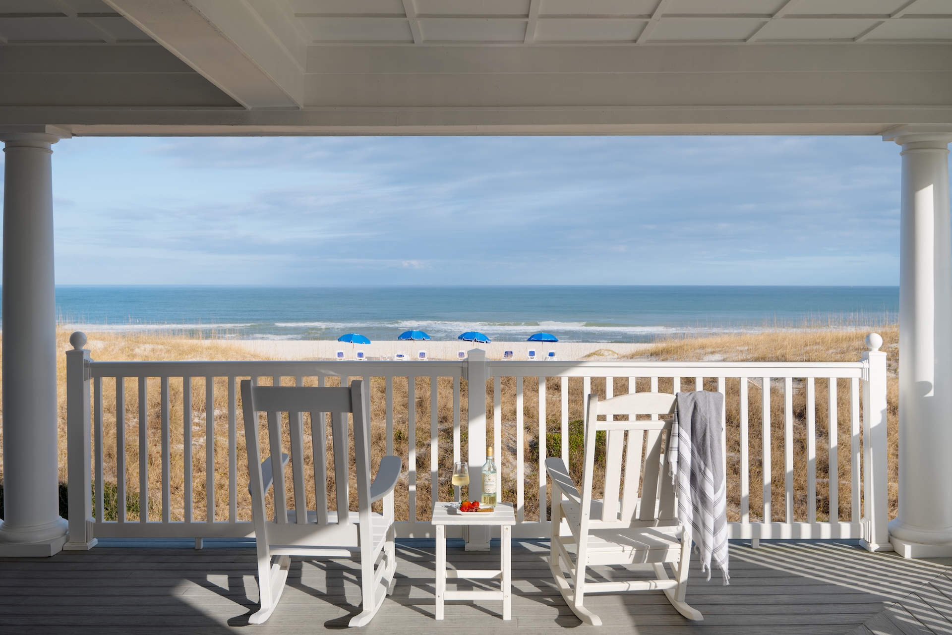 Two white rocking chairs and a small table sit on a porch overlooking sandy dunes and the ocean. Blue beach umbrellas line the shore under a partly cloudy sky. A towel hangs on one chair, and drinks rest on the table.