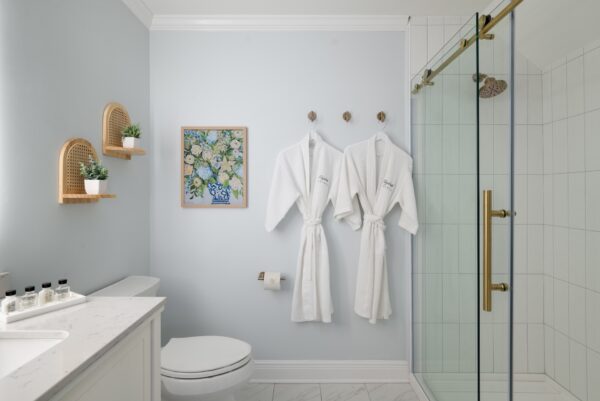 A modern bathroom with light blue walls, two white robes hanging on hooks, a glass shower with brass fixtures, a toilet, marble floor, and vanity. Two small shelves with potted plants and a floral painting are on the wall.