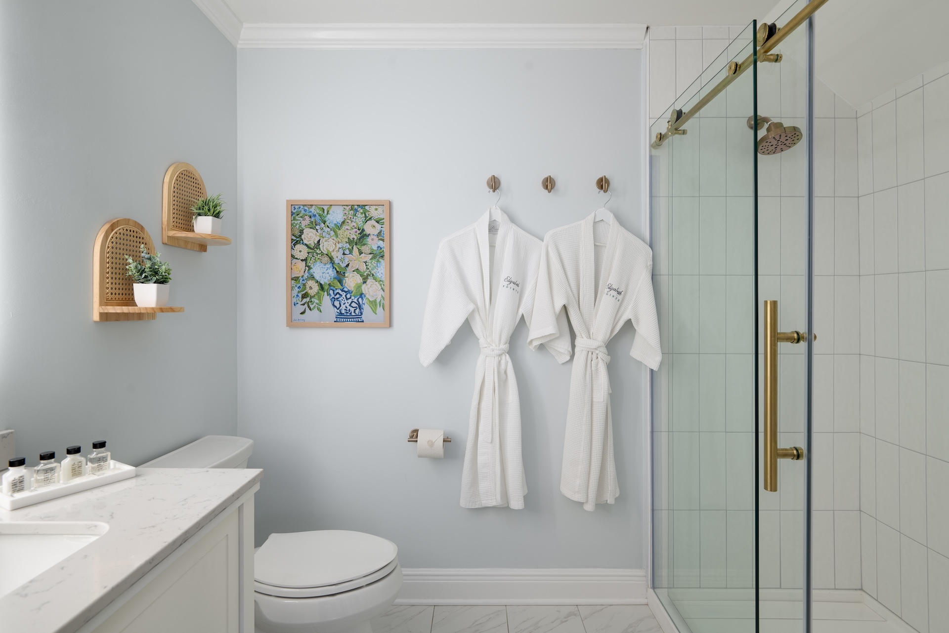 A modern bathroom with light blue walls, two white robes hanging on hooks, a glass shower with brass fixtures, a toilet, marble floor, and vanity. Two small shelves with potted plants and a floral painting are on the wall.