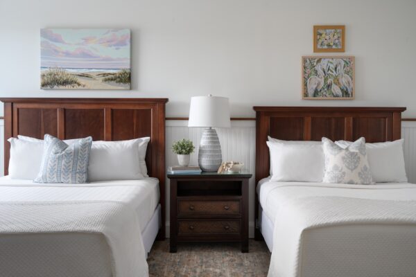 Two neatly made beds with wooden headboards are separated by a nightstand holding a lamp, plants, and decor. Three paintings hang on the white wall above the beds, creating a calm, inviting bedroom atmosphere.