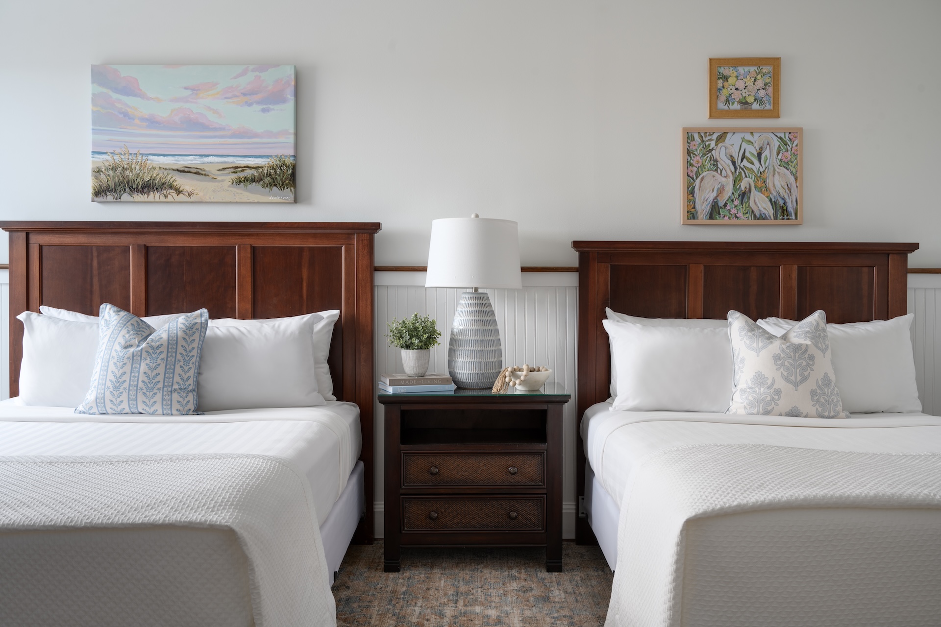 Two neatly made beds with wooden headboards are separated by a nightstand holding a lamp, plants, and decor. Three paintings hang on the white wall above the beds, creating a calm, inviting bedroom atmosphere.