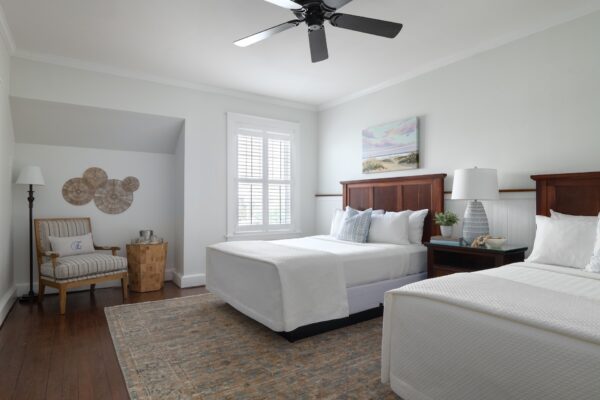 A bright bedroom with two double beds, white bedding, wooden headboards, a window with shutters, a ceiling fan, a nightstand with decor, and a cozy chair with a side table in the corner.