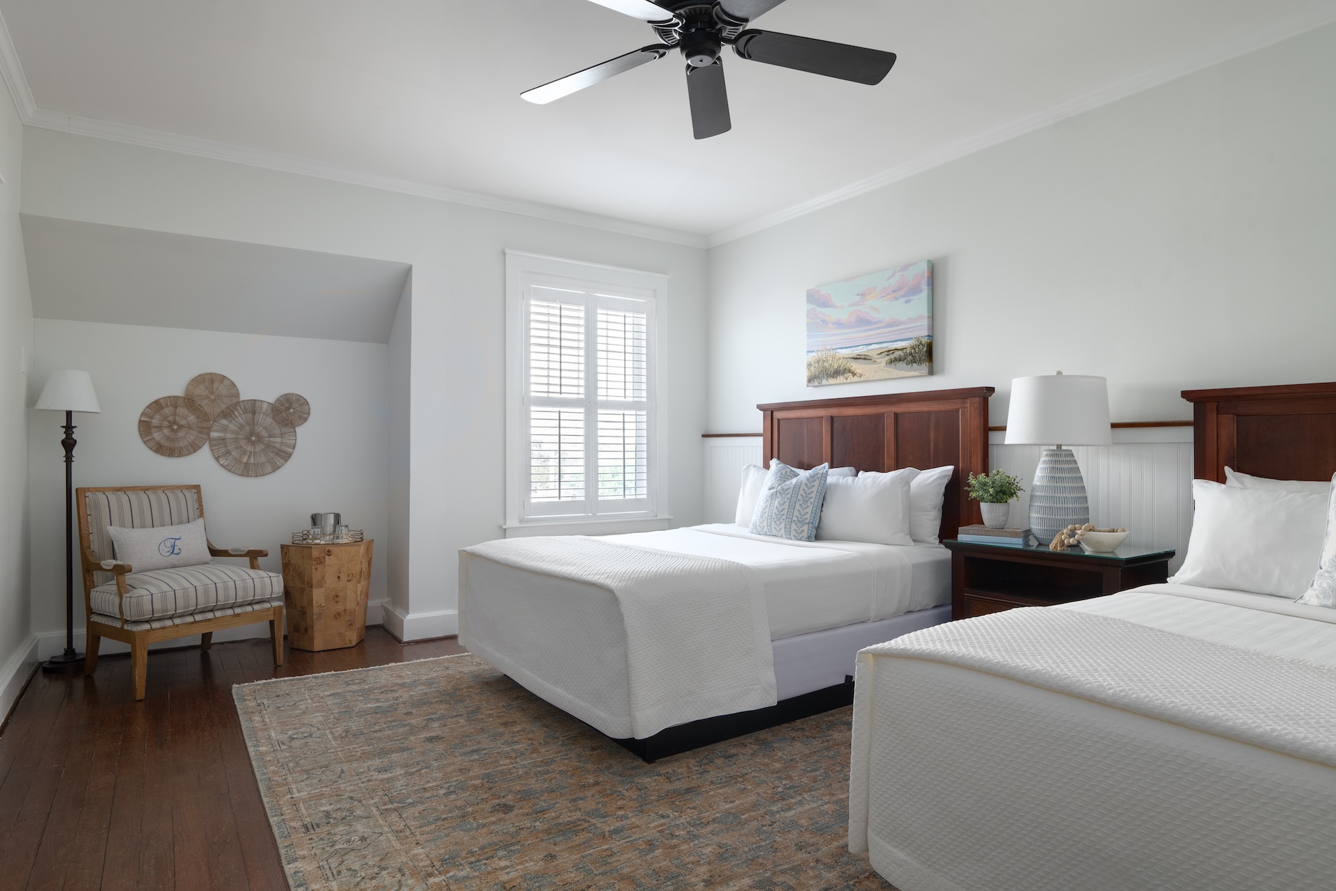 A bright bedroom with two double beds, white bedding, wooden headboards, a window with shutters, a ceiling fan, a nightstand with decor, and a cozy chair with a side table in the corner.