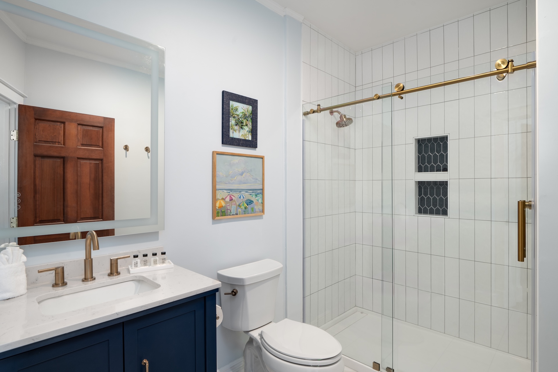 Modern bathroom with a glass shower, gold fixtures, a blue vanity with a white countertop, wall art above the toilet, and light blue walls. The shower has white tiles and a black tile accent.