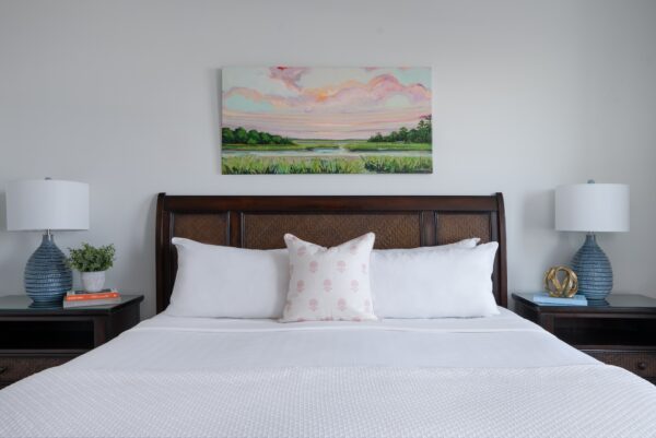 A neatly made bed with white bedding and a decorative pillow sits between two blue lamps on bedside tables at Elizabeth Pointe on Amelia Island. A colorful landscape painting hangs on the wall above the dark wooden headboard.