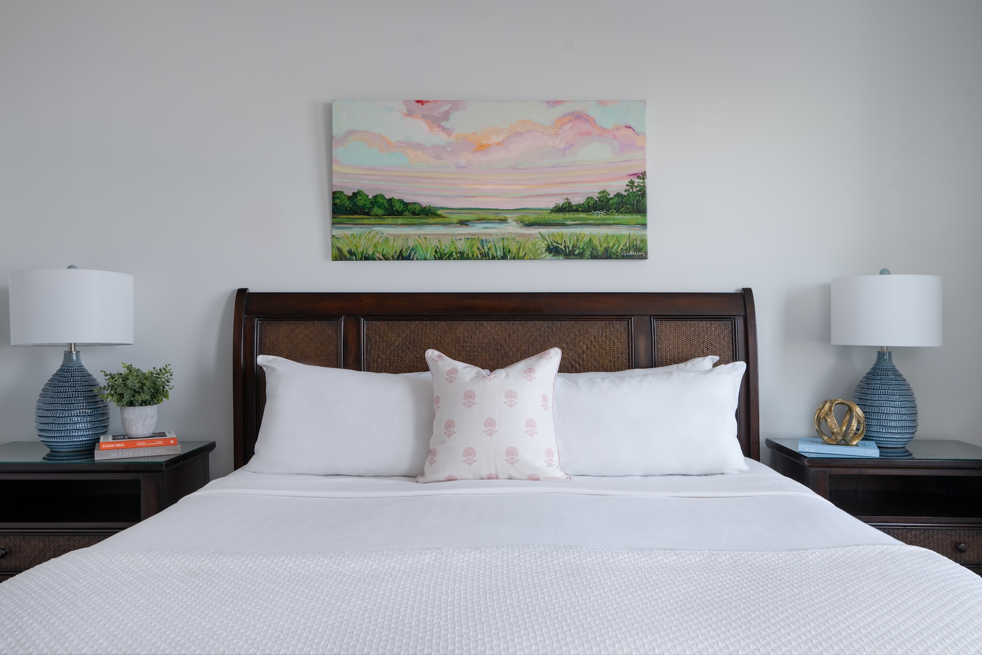 A neatly made bed with white bedding and a decorative pillow sits between two blue lamps on bedside tables at Elizabeth Pointe on Amelia Island. A colorful landscape painting hangs on the wall above the dark wooden headboard.