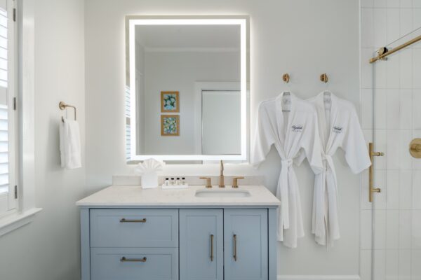A modern bathroom features a light blue vanity with a white countertop, an illuminated mirror, two white robes hanging on the wall, gold fixtures, a glass shower door, and framed art on the wall.