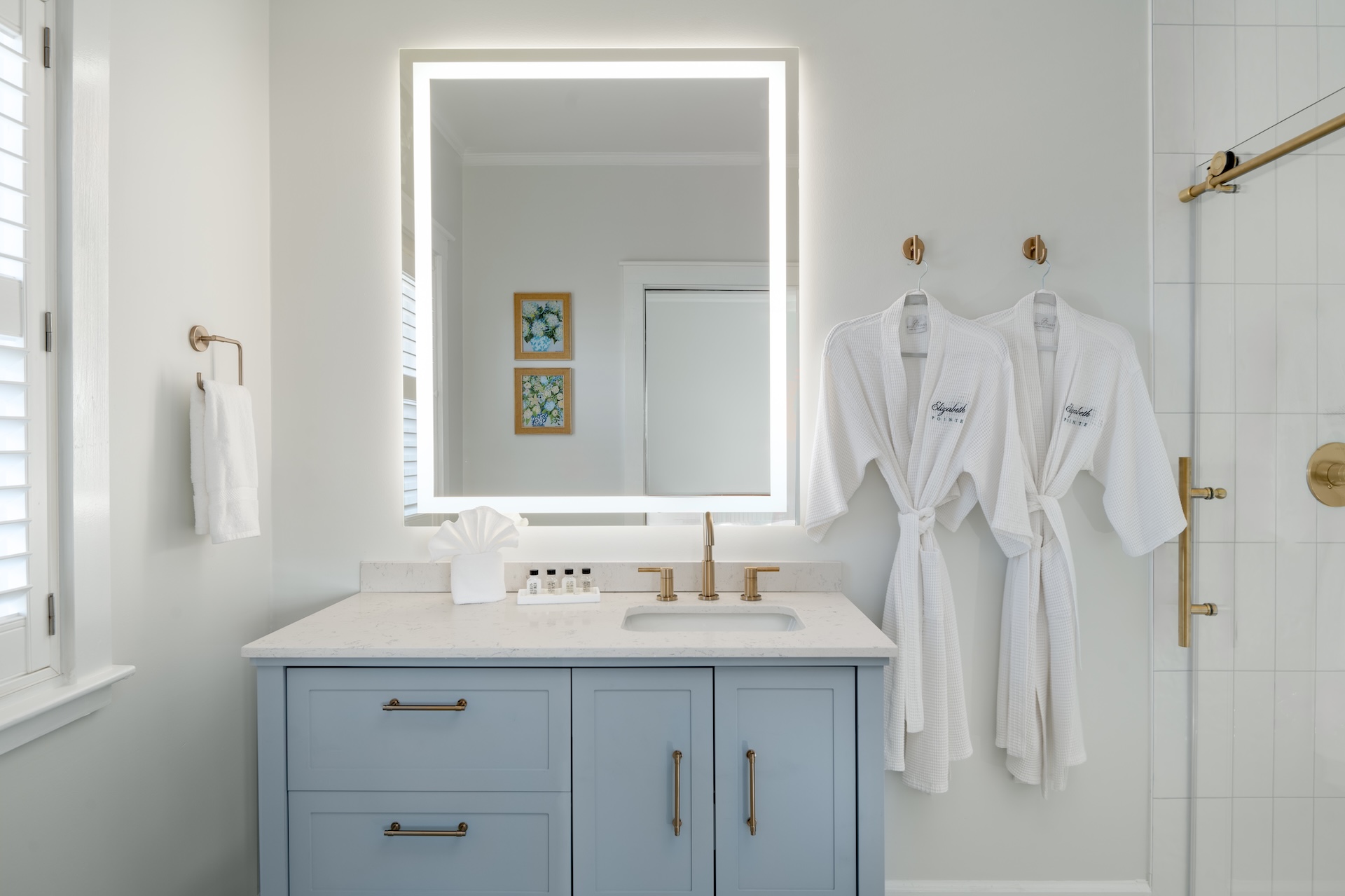 A modern bathroom features a light blue vanity with a white countertop, an illuminated mirror, two white robes hanging on the wall, gold fixtures, a glass shower door, and framed art on the wall.