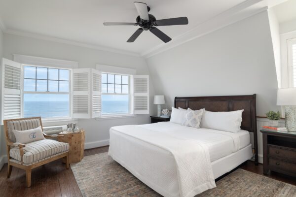 A bright bedroom at Elizabeth Pointe on Amelia Island features a large bed, two nightstands, a striped armchair, a wooden side table, and windows with white shutters overlooking the ocean. A black ceiling fan hangs above and natural light fills the room.