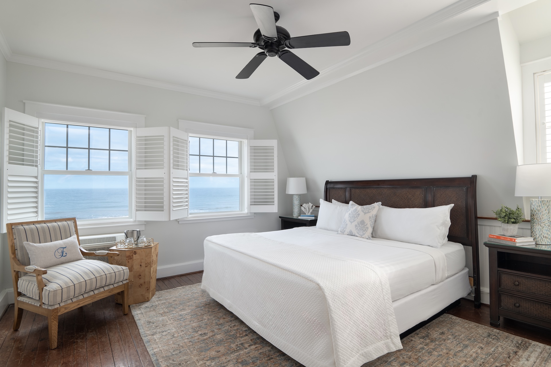 A bright bedroom at Elizabeth Pointe on Amelia Island features a large bed, two nightstands, a striped armchair, a wooden side table, and windows with white shutters overlooking the ocean. A black ceiling fan hangs above and natural light fills the room.