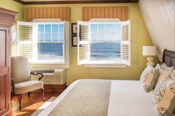 A cozy bedroom with yellow walls, a bed with patterned pillows, a wooden chair, and two windows with open shutters offering a scenic view of the ocean and blue sky. Sunlight brightens the room.