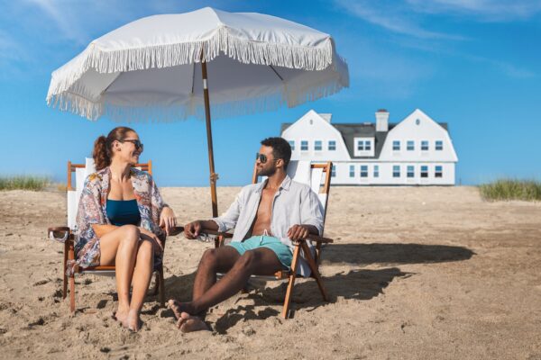 A woman and a man relax on beach chairs under a large white umbrella, smiling at each other. A white house with blue shutters sits in the background on a sunny day.