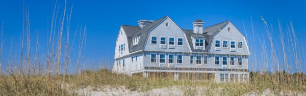 A large, white, two-story beach house with multiple windows and chimneys stands behind sand dunes and tall grass under a clear blue sky.