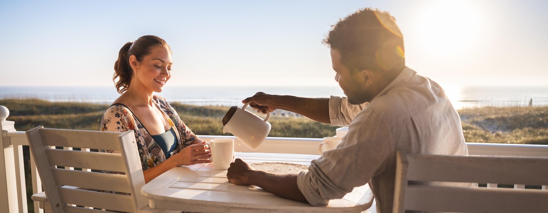 A man pours coffee into a womans mug as they sit at a table on a sunlit porch overlooking the beach and ocean during sunset.