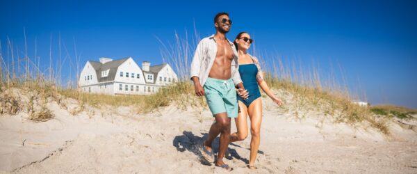 A smiling couple in swimwear holds hands while walking on a sandy beach with tall grass. A large white house with dormer windows is visible in the background under a clear blue sky.