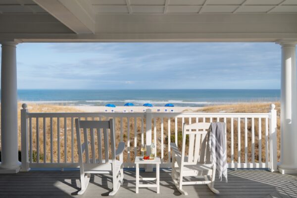 Two white rocking chairs and a small table with drinks sit on a porch overlooking a sandy beach with sea grass, blue umbrellas, and the ocean under a partly cloudy sky.