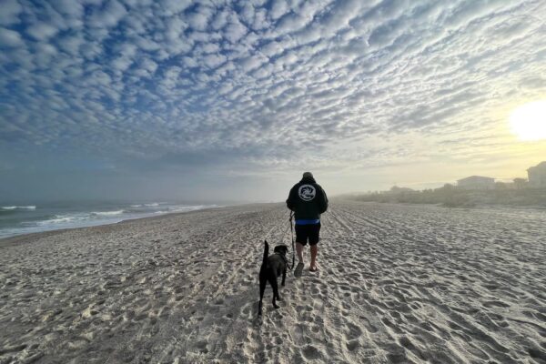 A person wearing a hoodie walks a black dog along a sandy beach under a dramatic sky filled with scattered clouds, with the sun low on the horizon.