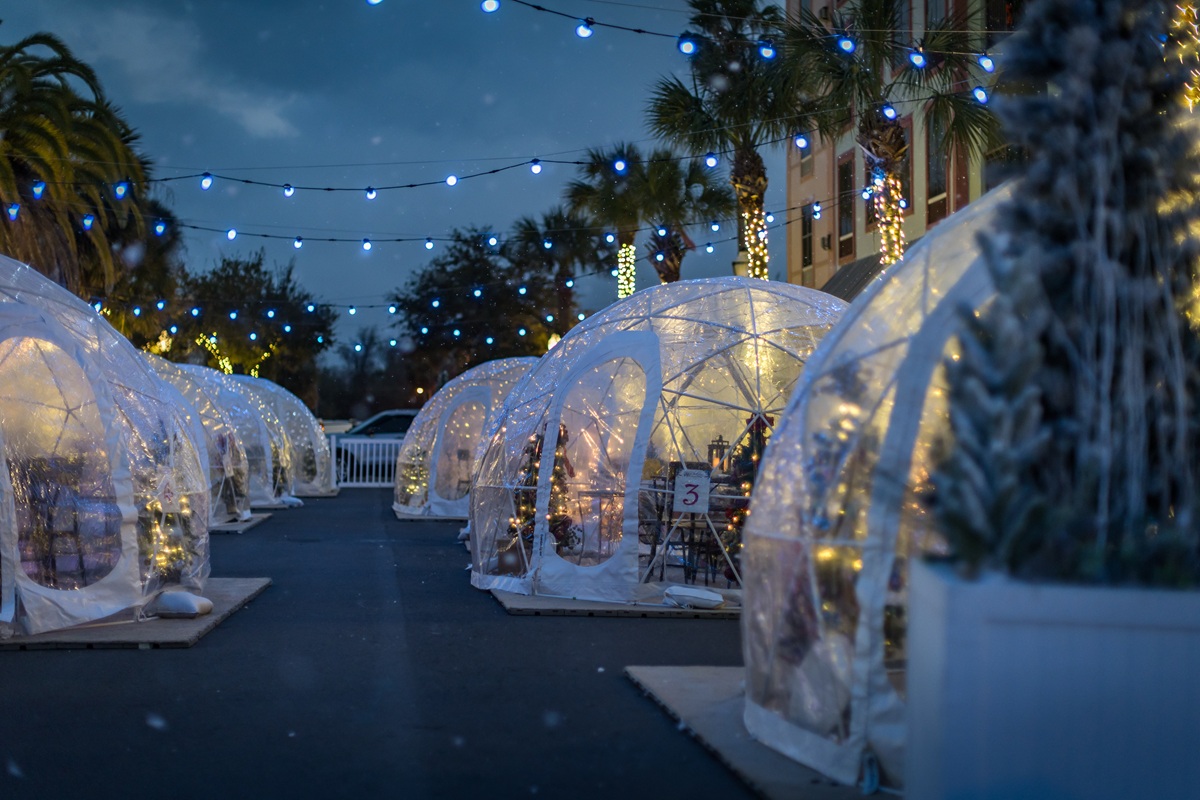 Outdoor dining domes with string lights are set up along a path at night. Each dome is lit warmly inside, surrounded by holiday decorations and palm trees wrapped in lights. Snow adds a festive, wintery touch.