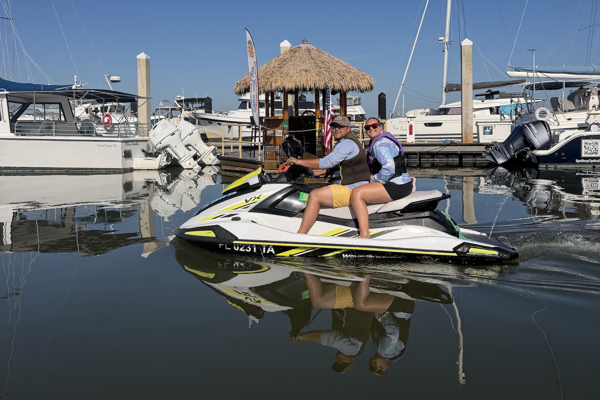 Two people wearing life jackets ride a jet ski on calm water in a marina, surrounded by docked boats and a small thatched-roof hut under a clear blue sky.