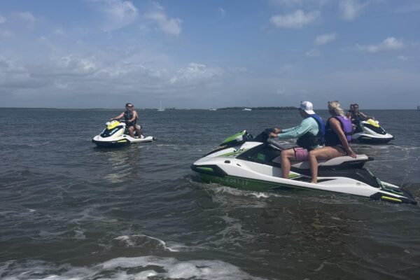 Three people on jet skis are riding on a large body of water under a partly cloudy sky, with two people on one jet ski and a single rider on another. Land and boats are visible in the distance.