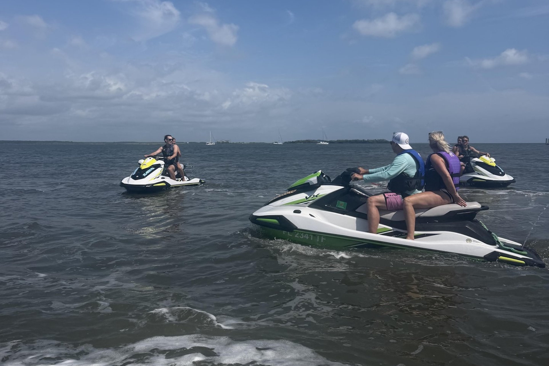 Three people on jet skis are riding on a large body of water under a partly cloudy sky, with two people on one jet ski and a single rider on another. Land and boats are visible in the distance.
