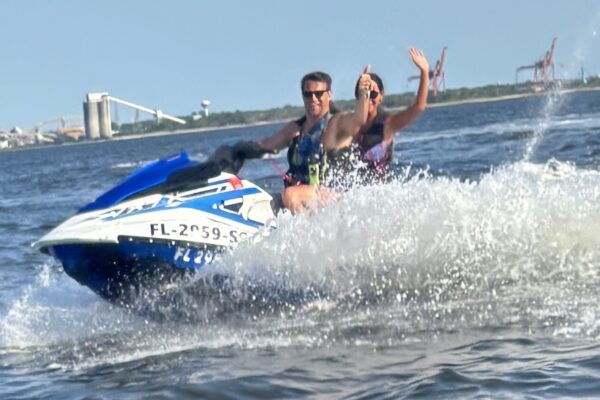Two people ride a blue and white jet ski on the water, both wearing life jackets. The person in front gives a thumbs up, while the person behind waves. Splashes of water surround them; industrial structures are visible in the background.