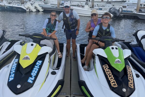Four people, two adults and two kids, smile on a dock with two colorful jet skis in front of them. They all wear life jackets and sunglasses, with boats and water in the background.