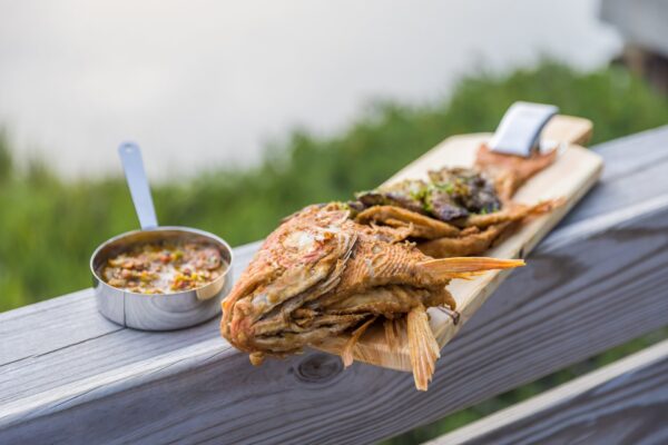 A whole fried fish served on a wooden board alongside a small metal bowl of chunky dipping sauce, with a blurred green outdoor background.