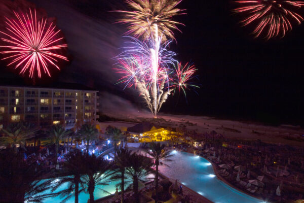 Fireworks light up the night sky over a brightly lit resort with palm trees, pools, and people gathered around, creating a festive atmosphere by the beach.