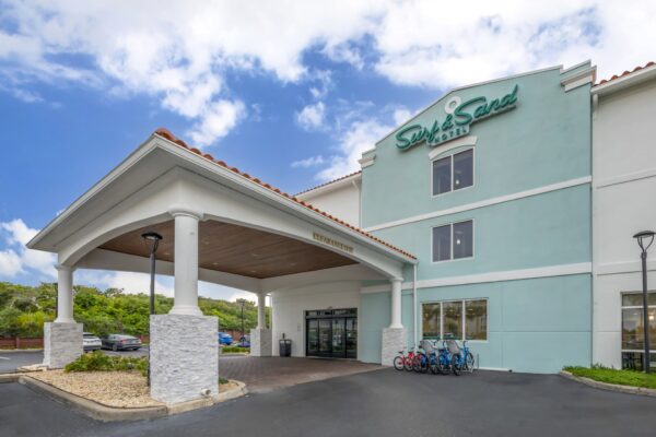 A light blue and white hotel called Surf & Sand with a covered entrance, glass doors, and several bicycles parked outside on a sunny day.