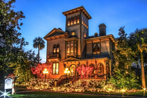 A large, ornate Victorian house with warm yellow lights and red holiday lights illuminating the front yard, surrounded by trees, at dusk with a clear blue sky.