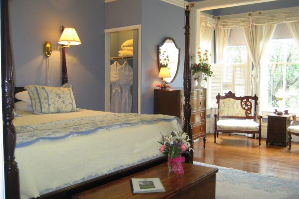 A cozy bedroom with a four-poster bed, yellow and blue bedding, flowers on a wooden chest, dresser with mirror, two armchairs by a bright bay window, and an open closet with neatly hung clothes and towels.