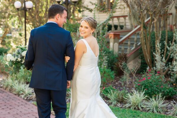 A bride in a white dress smiles back at the camera while holding hands and walking with a groom in a dark suit along a brick path in a garden setting.
